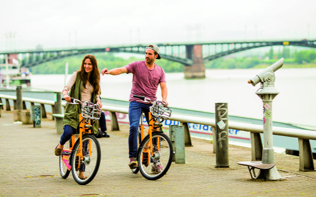 Zwei junge Menschen fahren mit Mieträdern am Rhein entlang 3